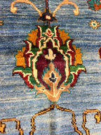 Macro close-up of a hand-knotted Sultani floral medallion on a Chobi Afghan carpet — deep burgundy, forest green, gold, ivory, and teal motif on a sky blue natural wool field, showcasing 35×40 knots per 10 cm artisan precision — Golden Loom Afghanistan, Jawzjan