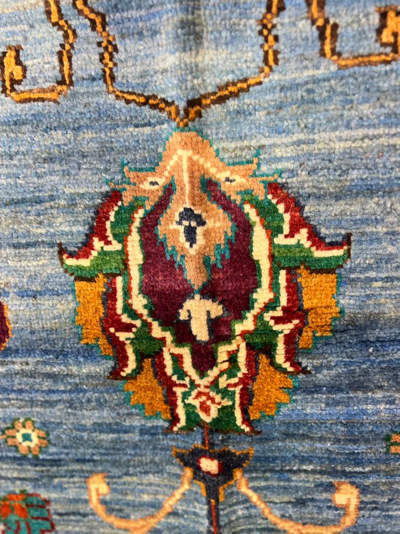 Macro close-up of a hand-knotted Sultani floral medallion on a Chobi Afghan carpet — deep burgundy, forest green, gold, ivory, and teal motif on a sky blue natural wool field, showcasing 35×40 knots per 10 cm artisan precision — Golden Loom Afghanistan, Jawzjan