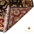 A close-up of an ornate Persian-style area rug with a dark navy/black field, red border, and cream floral motifs, shown with one corner folded over to reveal the underside and fringe. The Golden Loom Afghanistan logo is visible in the bottom right on a white background.