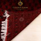 A close-up of a dark red and black patterned rug with white fringe, shown folded over on a light beige surface. The brand name 'Golden Loom Afghanistan' is printed at the top.