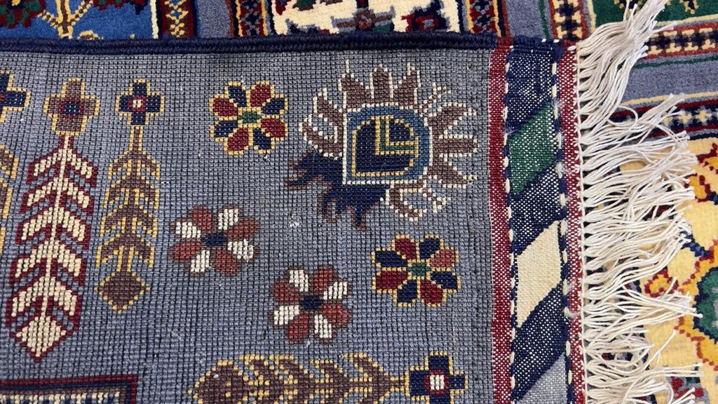 Belzhiki & Regional Afghan carpet close-up – hand-knotted wool pile detail showing individual knots, floral rosettes and botanical motifs in slate-blue, burgundy, gold and ivory, with hand-tied ivory fringe and striped border visible, 40×40 knots per 10cm, Qarqin, Jawzjan, Afghanistan