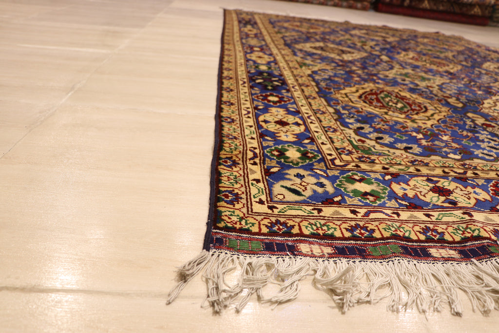 A detailed side angle view of a traditional Persian rug featuring intricate blue and yellow geometric patterns with white fringe tassels on a light-colored tile floor