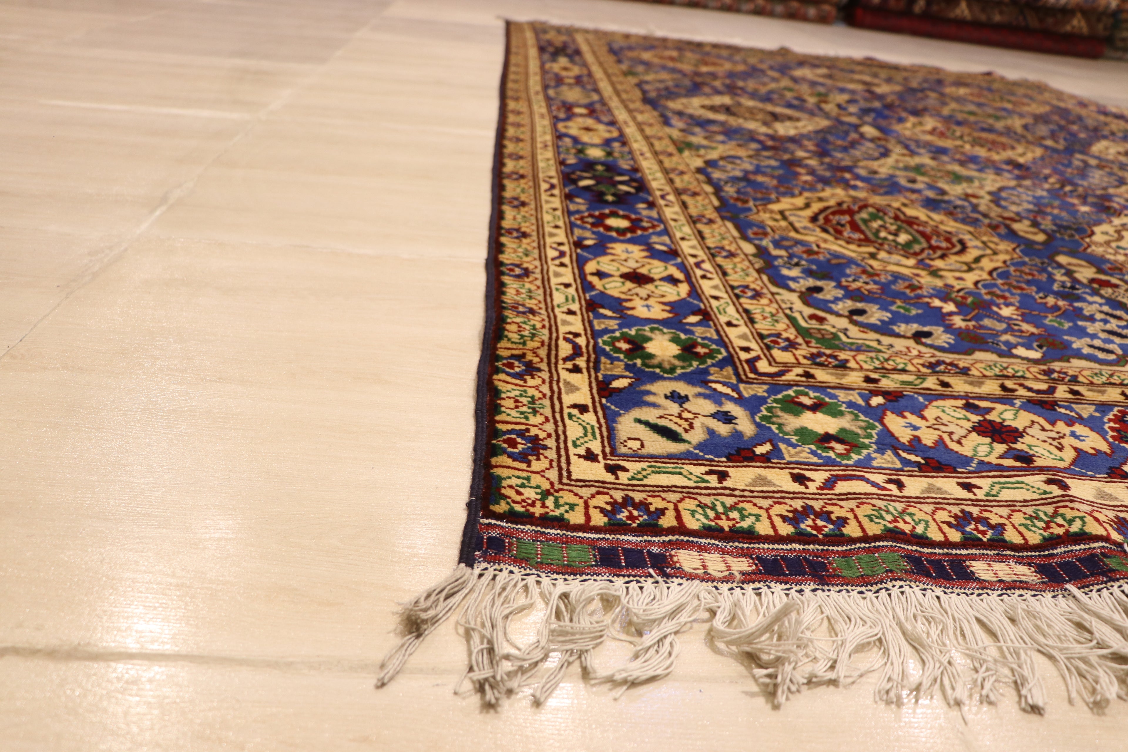 A detailed side angle view of a traditional Persian rug featuring intricate blue and yellow geometric patterns with white fringe tassels on a light-colored tile floor