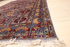 Corner view of a traditional blue-toned Persian rug with intricate geometric and floral patterns, featuring cream-colored fringe on a light-colored tile floor