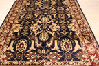 A traditional oriental area rug with an intricate floral and medallion pattern, featuring a dark navy blue center field, red borders, and beige and gold accents, laid out on a light wood floor.