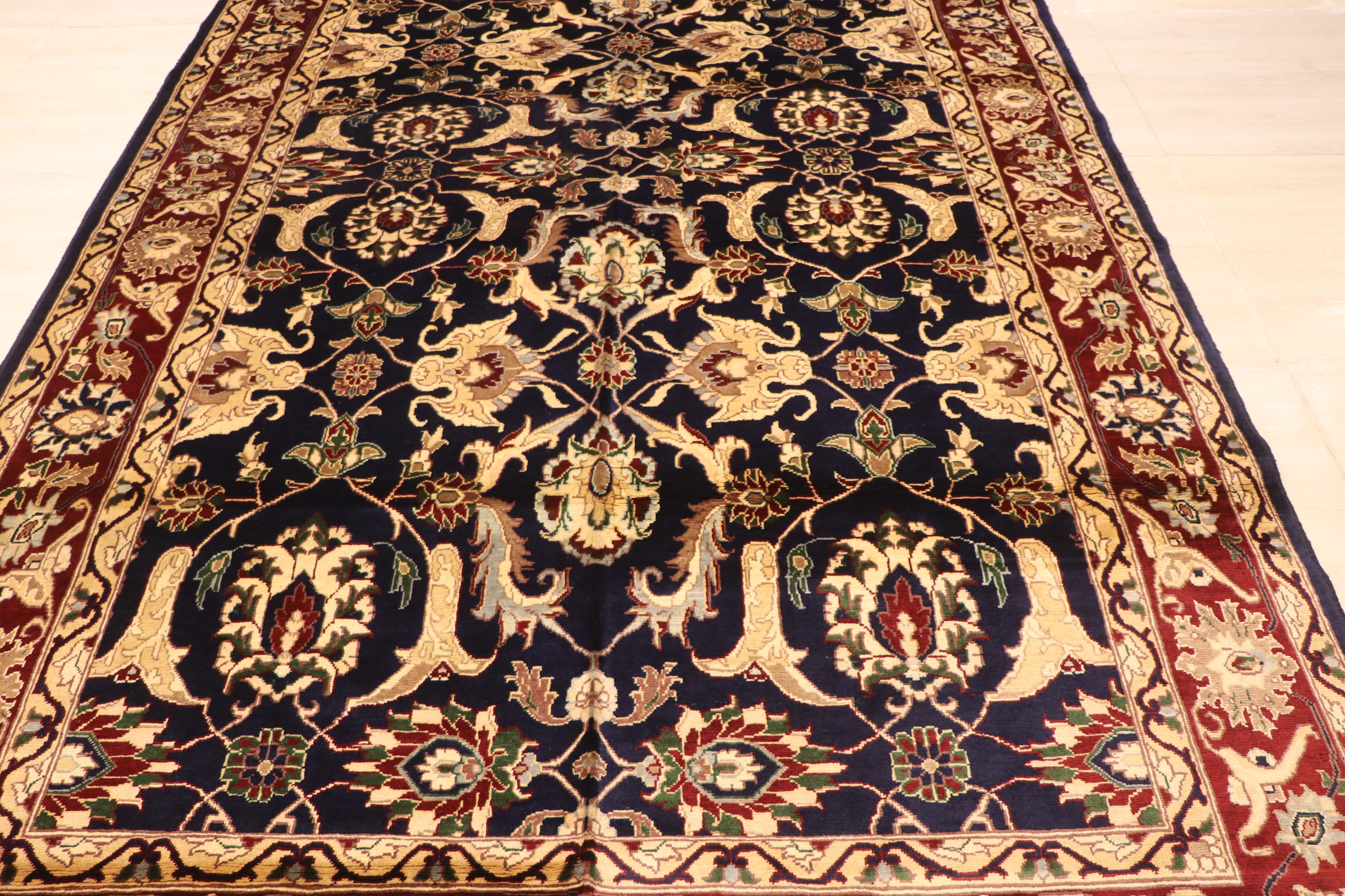 A traditional oriental area rug with an intricate floral and medallion pattern, featuring a dark navy blue center field, red borders, and beige and gold accents, laid out on a light wood floor.