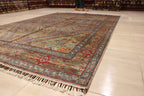Handwoven Afghan carpet displayed on showroom floor with multicolor floral border and tassel fringe, surrounded by rolled rugs