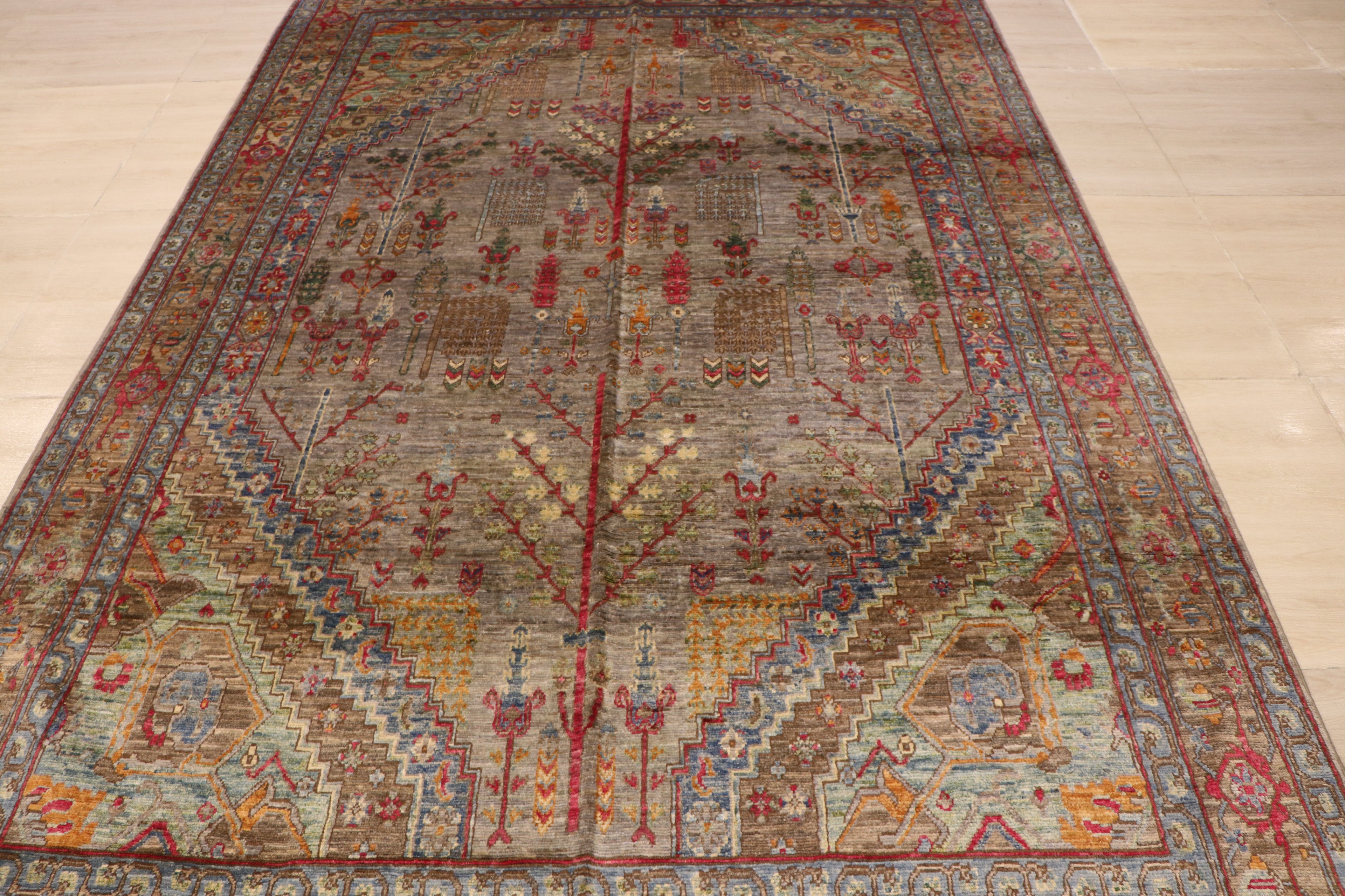 Close-up of handwoven Afghan rug featuring symmetrical tree-of-life and floral motifs on a warm grey field with crimson and gold accents