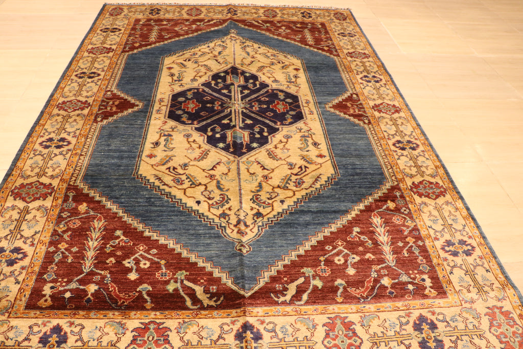 A traditional oriental area rug with a geometric medallion pattern, featuring a blue-gray center, red corners, and beige and gold borders, laid out on a light wood floor.