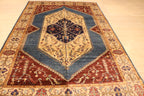 A traditional oriental area rug with a geometric medallion pattern, featuring a blue-gray center, red corners, and beige and gold borders, laid out on a light wood floor.