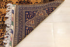 A close-up of a folded traditional patterned rug showing its cream fringe, navy blue border, and warm tan field on a light floor.