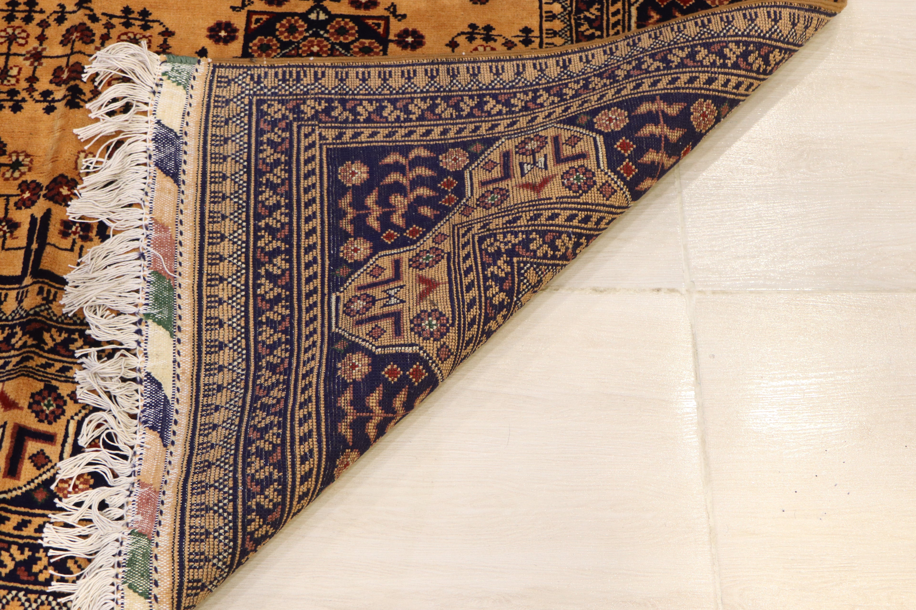 A close-up of a folded traditional patterned rug showing its cream fringe, navy blue border, and warm tan field on a light floor.