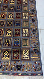 Belzhiki & Regional Mirror Design Afghan carpet photographed on white tile floor – angled view showing botanical panel grid with cypress trees, rosettes and floral motifs in slate-blue, burgundy, gold, ivory and green, decorative border with arrow and floral motifs, ivory fringe at bottom, 202×301cm, 100% Turkman wool, Qarqin, Jawzjan, Afghanistan