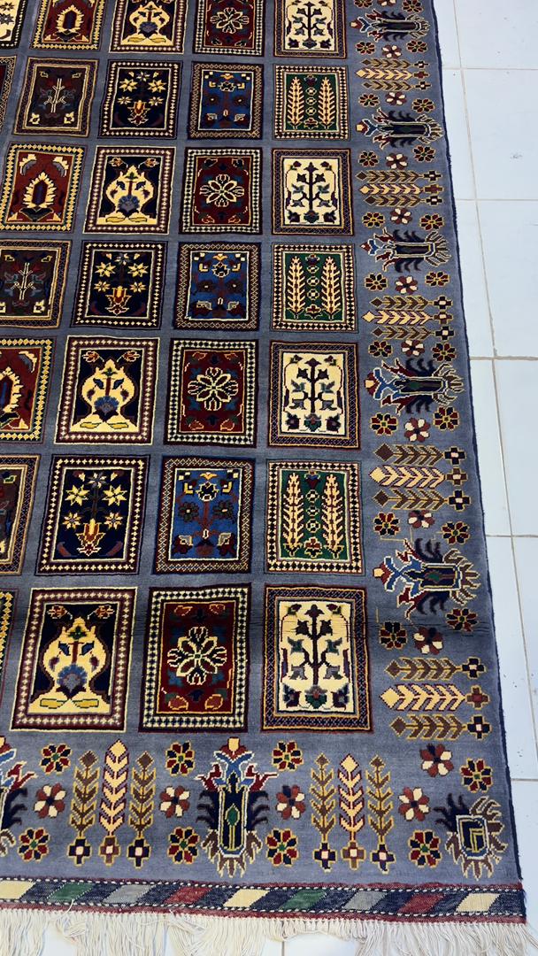 Belzhiki & Regional Mirror Design Afghan carpet photographed on white tile floor – angled view showing botanical panel grid with cypress trees, rosettes and floral motifs in slate-blue, burgundy, gold, ivory and green, decorative border with arrow and floral motifs, ivory fringe at bottom, 202×301cm, 100% Turkman wool, Qarqin, Jawzjan, Afghanistan